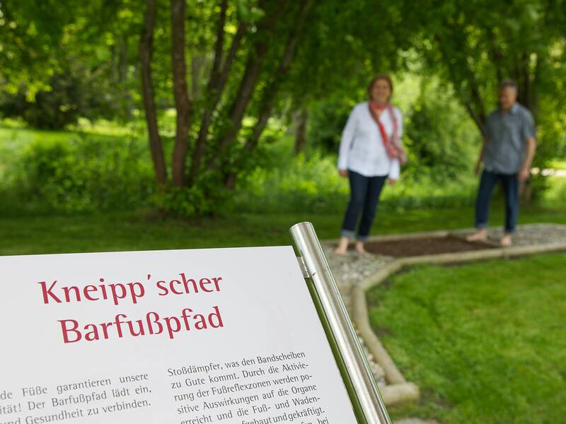 Schild mit rotem Text 'Kneipp'scher Barfußpfad' und Erläuterungstext, im Hintergrund zwei Personen auf einem Barfußpfad im Park.
