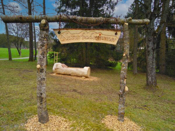 Holztor aus Baumstämmen mit hölzernem Schild 'Naturbadezimmer' in einer Wiese, im Hintergrund Baumstamm als Badewanne.