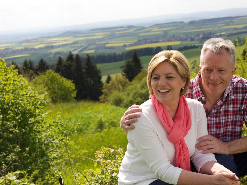 Älteres Paar sitzt auf einer Wiese mit Blick auf eine hügelige Landschaft und Felder im Hintergrund.