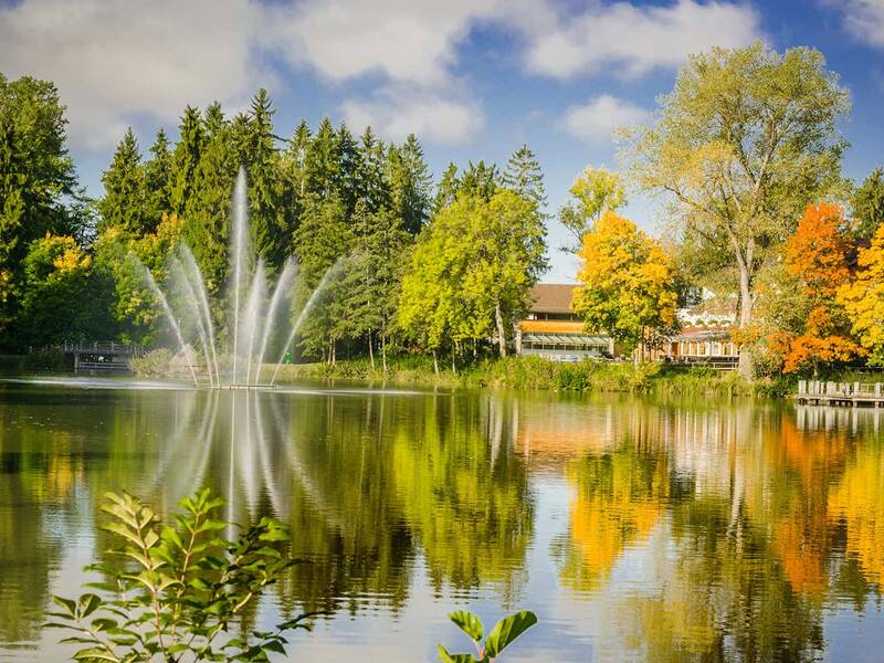 See mit Springbrunnen, umgeben von Bäumen mit grünen und herbstlich gefärbten Blättern, im Hintergrund das Hotel Salinensee