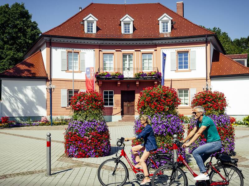Zwei Personen fahren mit Fahrrädern auf einem gepflastertem Platz vor dem Bad Dürrheimer Rathaus, einem zweistöckigen Gebäude mit roten Dachziegeln und Blumenschmuck an Fenstern und in großen Pflanzkübeln.