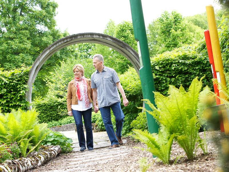 Älteres Paar geht auf einem Kiesweg durch einen Garten mit Farnen und bunten Stelen, im Hintergrund ein Metallbogen.
