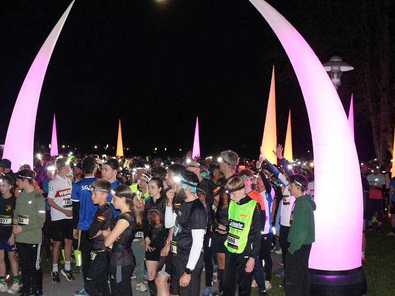 Gruppe von Läufern in Sportkleidung und Stirnlampe bei Nacht vor großen, leuchtenden, spitz zulaufenden Säulen im Freien.