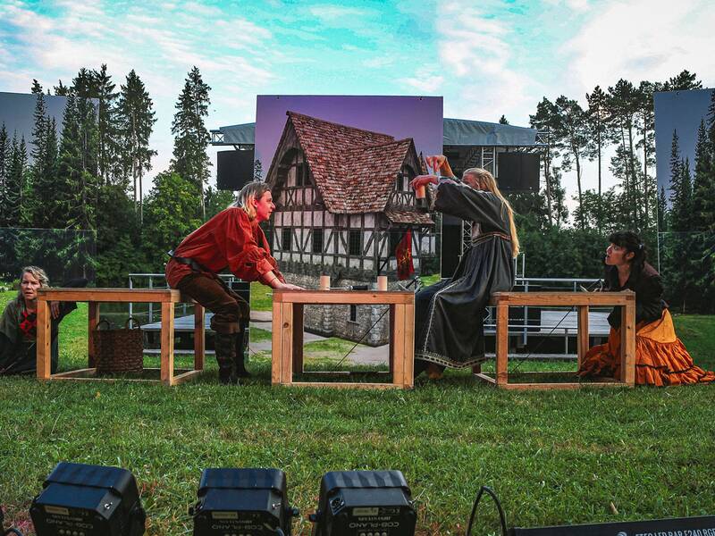 Vier Personen in historischer Kleidung sitzen an einem Holztisch auf einer Wiese vor einer Kulisse mit Fachwerkhaus und Wald.