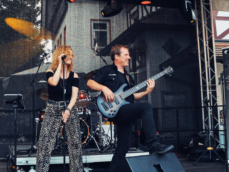 Zwei Musiker auf Bühne: Frau mit Mikrofon und Mann mit E-Bass, im Hintergrund Schlagzeug und Banner mit Schriftzug 'KARABOOM'.