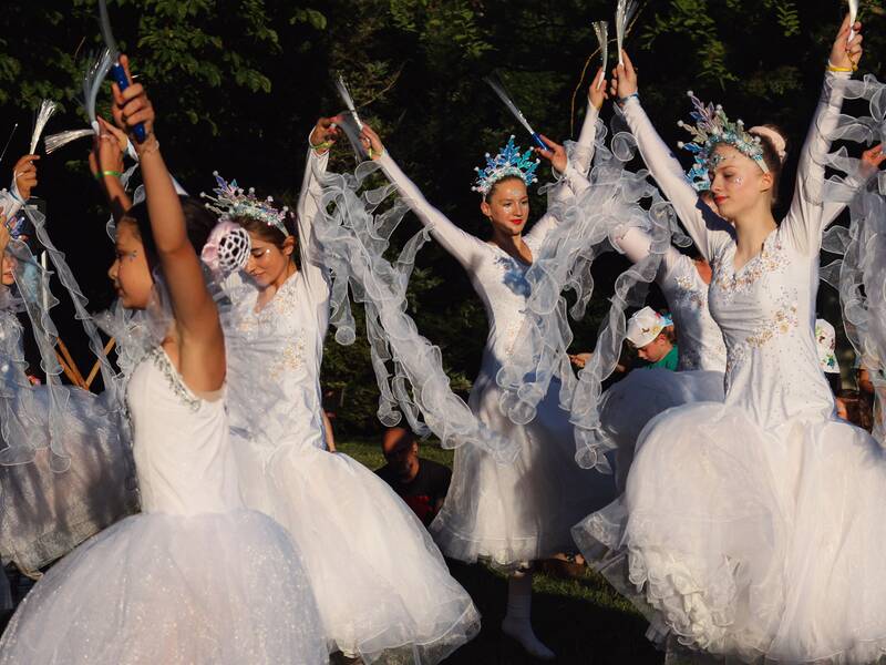 Mehrere Balletttänzerinnen in weißen Kleidern mit langen, transparenten Ärmeln und blauen Kopfschmuck heben Arme mit Leuchtstäben in einem Park..