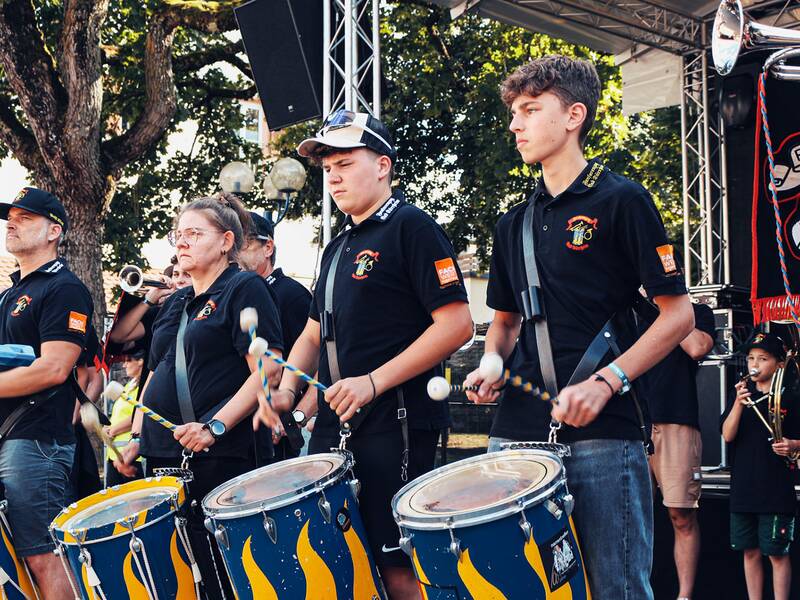 Fünf Musiker in schwarzen T-Shirts mit orangefarbenen Ärmeln spielen Trommeln und Fanfaren auf einer Bühne bei Tageslicht.