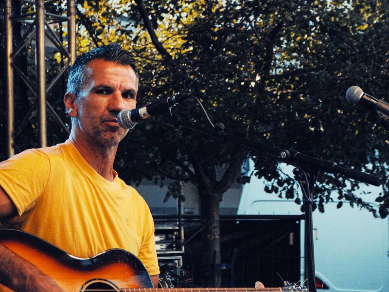 Mann in gelbem T-Shirt spielt Gitarre auf einer Bühne mit Mikrofonständern und Bäumen im Hintergrund.