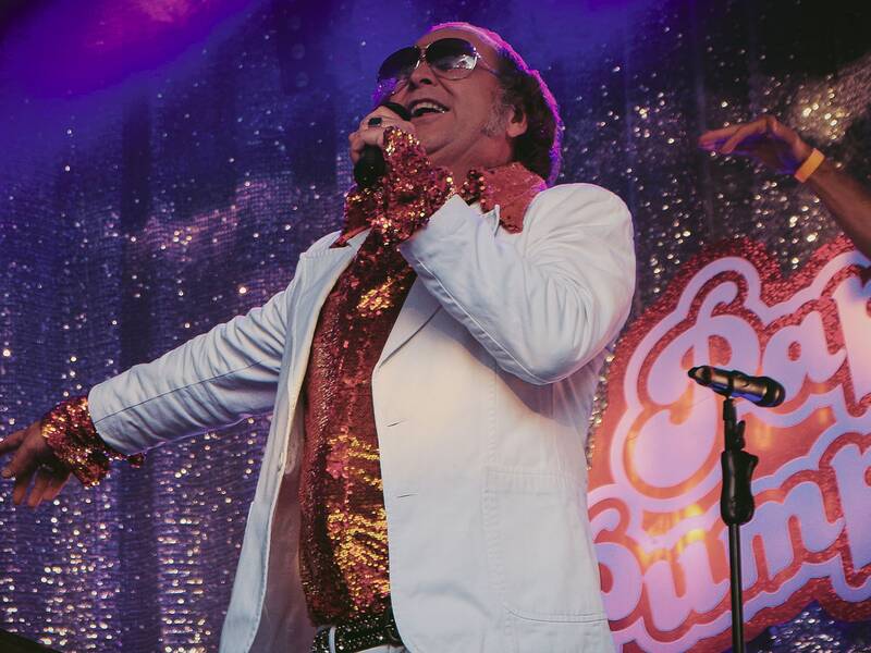 Person in weißem Sakko und glitzerndem, rot-goldenem Hemd singt auf Bühne vor lila-glitzerndem Hintergrund mit Schriftzug 'Papis Pumpels'.