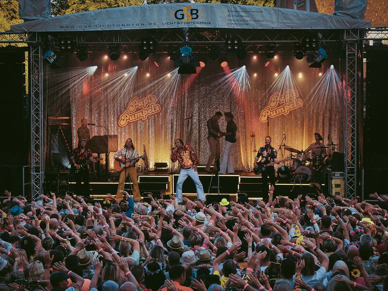 Bühne mit Band und Sänger vor großem Publikum, das Hände in die Luft streckt, Abendlicht und Bühnenbeleuchtung