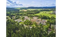Luftaufnahme eines Gebäudekomplexes mit mehreren flachen, rechteckigen Gebäuden, umgeben von Wiesen und Wald.