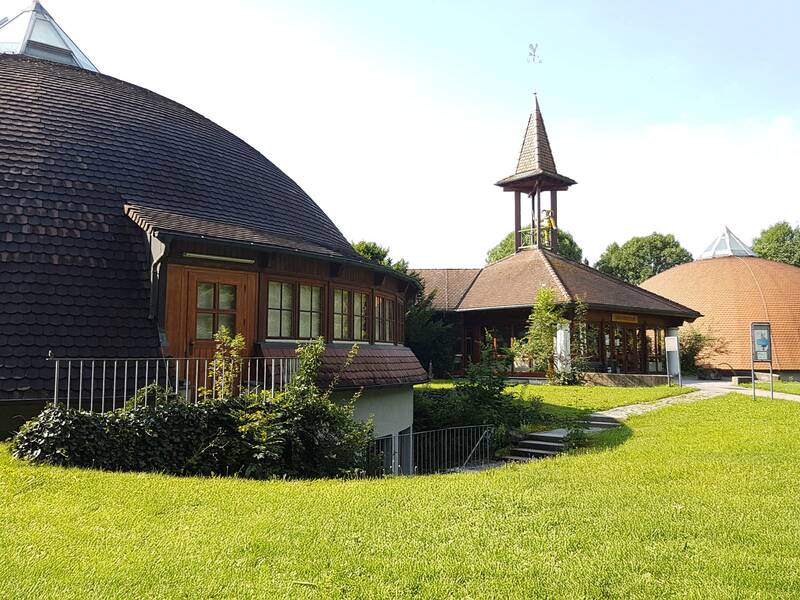 Gebäude mit kuppelförmigen Dächern und einem kleinen Turm mit spitzem Dach in einer grünen Parkanlage