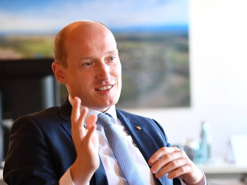 Bürgermeister Jonathan Berggötz sitzt in seinem Büro im Hintergrund sieht man ein Luftbild und einen Bildschirm