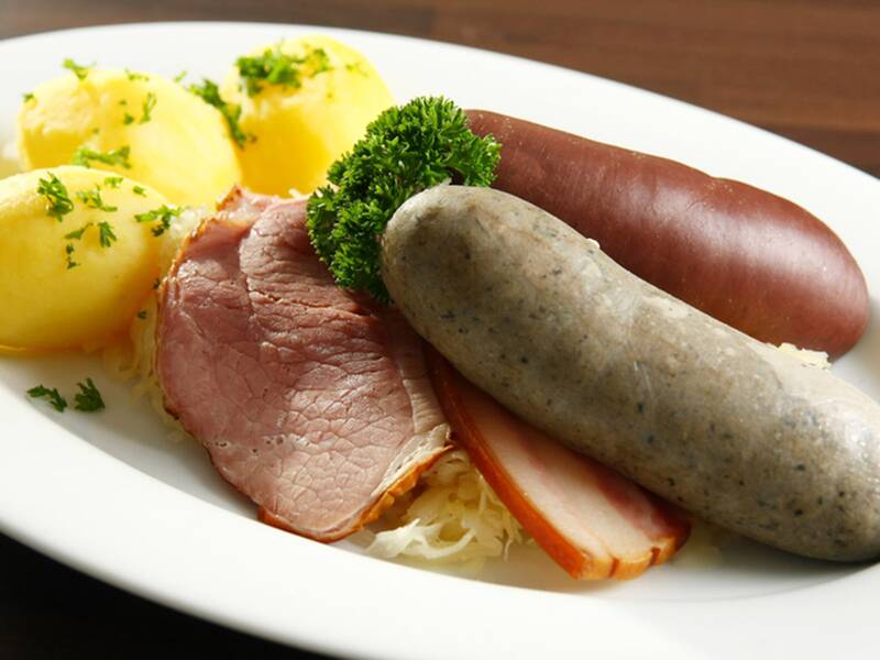 Teller mit kleinen Kartoffeln, zwei verschiedenen Würsten, Scheiben von geräuchertem Fleisch, Sauerkraut und Petersilienzweig