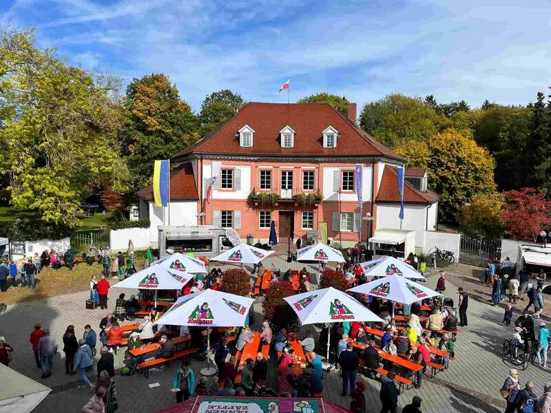 Marktplatz mit orangefarbenen Bänken und Tischen unter weißen Sonnenschirmen vor einem zweistöckigen Gebäude mit rotem Dach und Fahne, umgeben von herbstlich gefärbten Bäumen und zahlreichen Menschen.