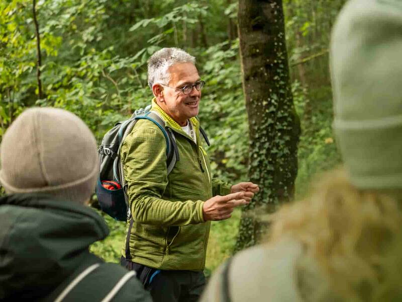 Ein Mann in grüner Jacke und mit grauen Haaren spricht im Wald zu einer Gruppe 