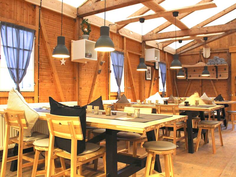 Innenraum einer Holzhütte mit Tischen, Stühlen, Hockern, hängenden Lampen und Fenstern mit blauen Vorhängen