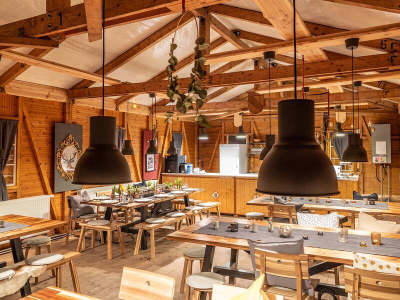 Innenansicht einer rustikalen Holzhütte mit gedeckten Tischen, Holzbalken und großen schwarzen Hängeleuchten
