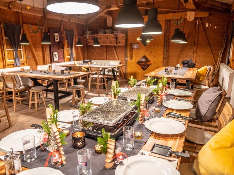 Rustikale Holzhütte mit langen Tischen, gedeckten Tellern, Raclette-Grillplatten und weihnachtlicher Dekoration