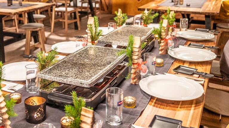 Rustikale Holzhütte mit langen Tischen, gedeckten Tellern, Raclette-Grillplatten und weihnachtlicher Dekoration