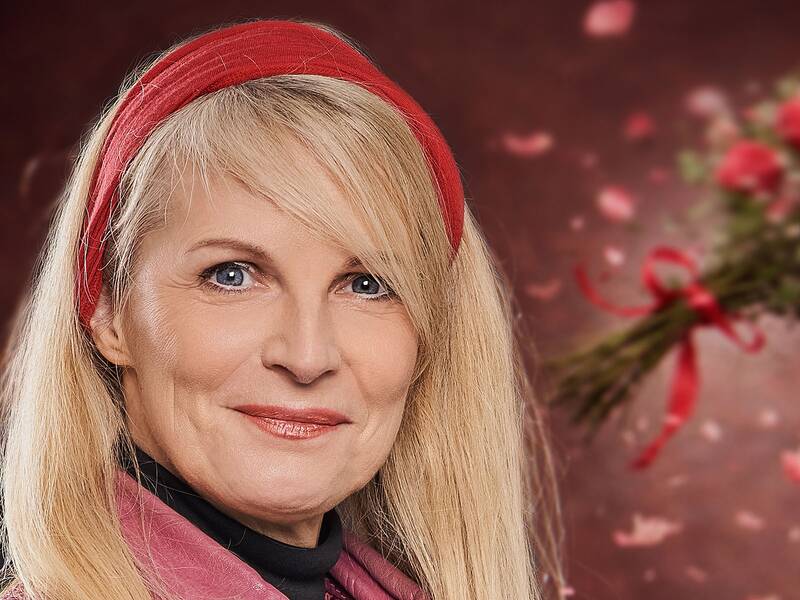 Frau mit langen blonden Haaren trägt ein rotes Stirnband, eine pinke Lederjacke und einen pinken Schal vor einem unscharfen Hintergrund mit einem Blumenstrauß