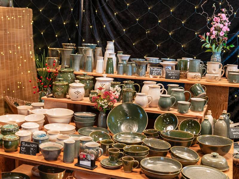 Tisch mit vielfältigem grünen Keramikgeschirr und Vasen auf einem Kunsthandwerkmarkt mit Lichterkette im Hintergrund