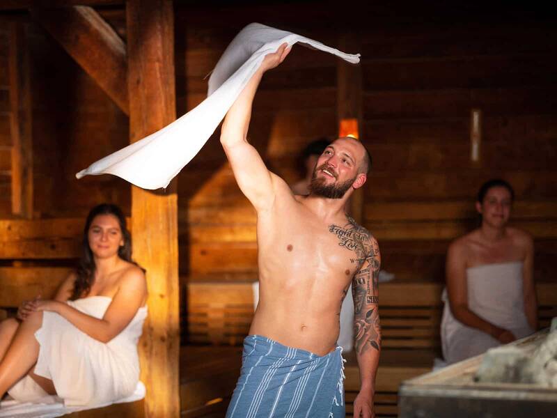 Saunemeister wedelt Handtuch in der Sauna. Personen sitzen auf den Bänken der Sauna.