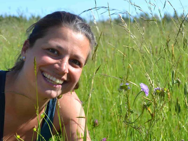 Lächelnde Frau mit einem braunen Pferdeschwanz sitzt in der Hocke in einer blühenden Wiese
