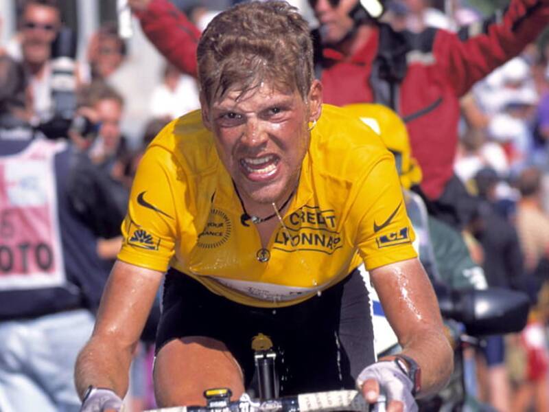Jan Ullrich in einem gelben Trikot der sehr angestengt auf einem Fahrrad sitzt. Im Hintergrund feuern ihn Fans an.