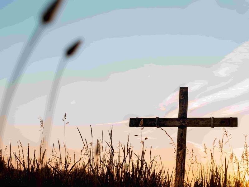 Ein Holzkreuz steht auf einer Wiese. Im Hintergrund geht die Sonne auf.
