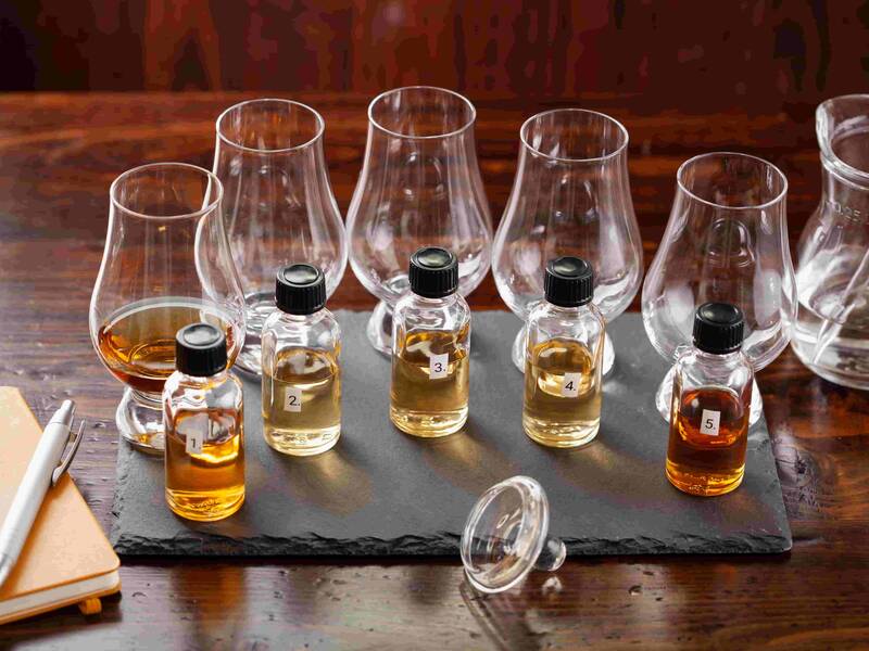 Sechs kleine Fläschchen mit nummerierten Etiketten und unterschiedlich gefärbten Whiskys stehen auf einer schwarzen Schieferplatte vor sechs leeren Whiskygläsern auf einem Holztisch.