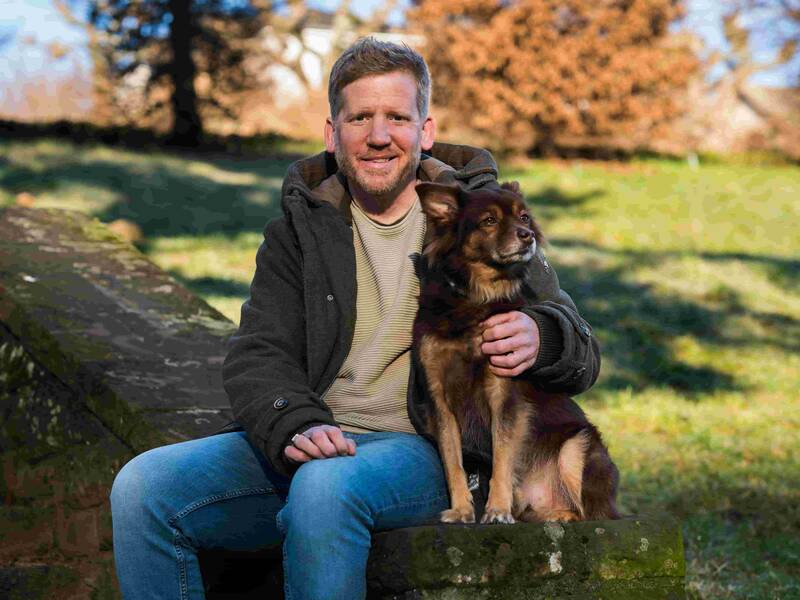 Männlich gelesene Person mit braunem Hund sitzt auf einer Steinmauer im Freien bei sonnigem Herbstwetter