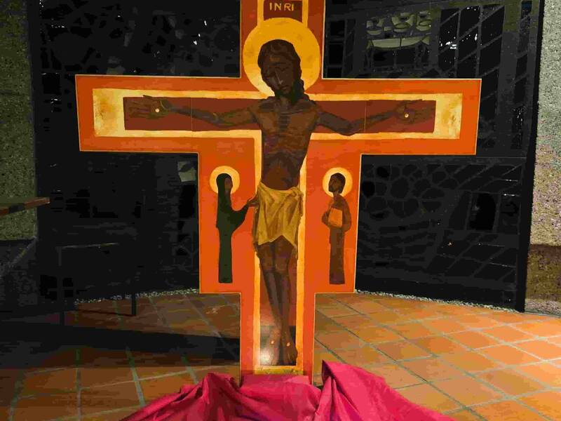 Kruzifix mit stilisierter Darstellung von Jesus vor rotem Tuch mit mehreren kleinen Kerzen auf Bodenfliesen in dunklem Raum