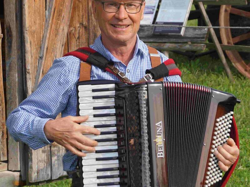 Mann mit Schiebermütze und Brille spielt Akkordeon vor einer Holzhütte im Freien