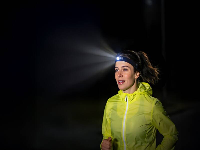 KI generiert: Frau in gelber reflektierender Laufjacke mit Stirnlampe, die einen Lichtstrahl in dunkler Umgebung aussendet.